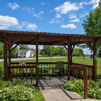 A wooden gazebo with outdoor furniture, including chairs and a table, set in a grassy area under a blue sky with scattered clouds, surrounded by lush green trees and plants.