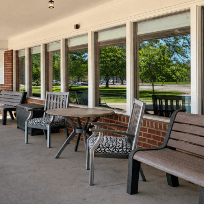 Outdoor patio with wooden and metal chairs and tables, overlooking a grassy area with trees through large glass windows.