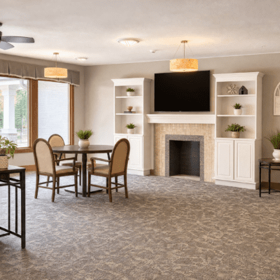 A spacious living room with a fireplace and built-in white shelves on either side, a large flat-screen TV above the fireplace, a round dining table with four chairs, and windows letting in natural light.