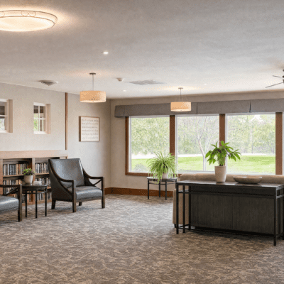 A spacious living room with large windows showing green outdoor scenery, furnished with a black armchair, a small wooden side table, a black console table with potted plants, and a bookshelf. The room has beige carpet, white walls, and ceiling lights.