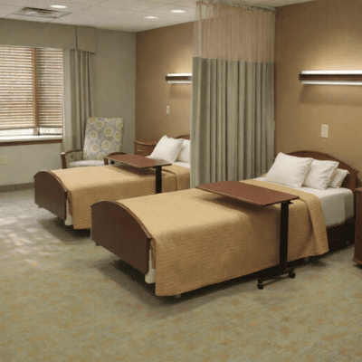 Two hospital beds with beige bedding and white pillows are positioned side by side in a room. Each bed has a wooden overbed table. There is a window with blinds, a patterned armchair, and a privacy curtain between the beds. The walls are beige, and there are wall-mounted lights above each bed.