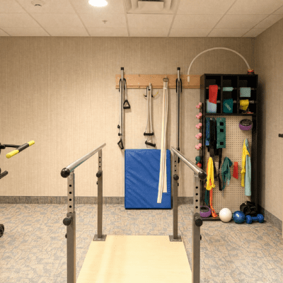 A therapy or exercise room with parallel bars in the foreground, padded mats, and various fitness accessories such as balls, resistance bands, and dumbbells organized on shelves and hooks against the wall.
