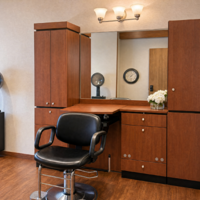 A hair salon station with a large mirror, wooden cabinets, a black salon chair, a globe, a clock, and a vase of white flowers on the countertop.