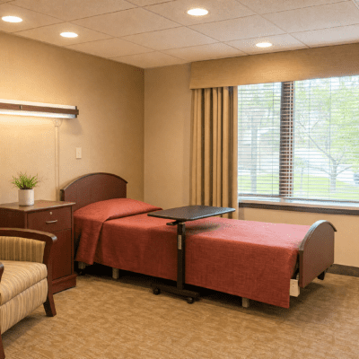 A bedroom with a single bed covered in a reddish-brown bedspread, a wooden headboard, and a small bedside table with a potted plant. There is a window with beige curtains, and a view of green trees outside. A tray table is positioned over the bed, and part of a striped armchair is visible on the left. The room has beige walls and carpet, with ceiling lights.