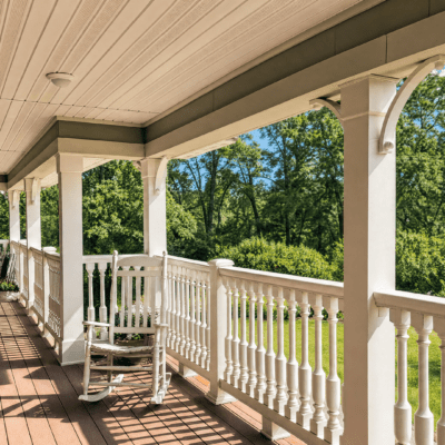 A covered porch with white railings and columns, overlooking a lush green yard with trees and grass on a sunny day.