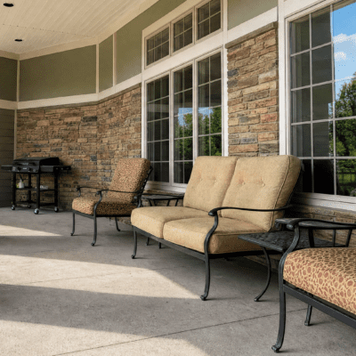 A covered outdoor porch with a beige cushioned sofa, two upholstered chairs, and a small black side table, with large windows and a stone wall in the background.