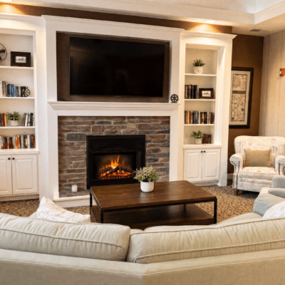 Living room with a large flat-screen TV mounted above a brick fireplace, flanked by built-in white bookshelves with decorative items and books; a wooden coffee table with a potted plant in front of a cream-colored sectional sofa and a patterned armchair.