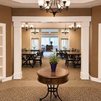 A dining area viewed through a large white-framed doorway, with a round wooden table holding a potted green plant in a purple pot in the foreground. The room has multiple tables and chairs, beige walls, and large windows with sheer curtains, illuminated by chandeliers and wall sconces.