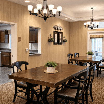A dining room with two wooden tables, each surrounded by black chairs. The walls are beige with framed pictures and decorative plates. There are two chandeliers hanging from the ceiling, and a window with curtains in the background. Potted plants are on the tables.