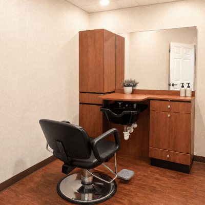 A small salon or barbershop station with a black barber chair in front of a wooden counter and cabinet, a mirror on the wall, and a sink with a potted plant on the counter. The room has beige walls and wooden flooring.